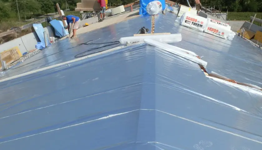 Lavoratori installano un freno vapore su un tetto in costruzione, con rotoli di materiale isolante in primo piano.
