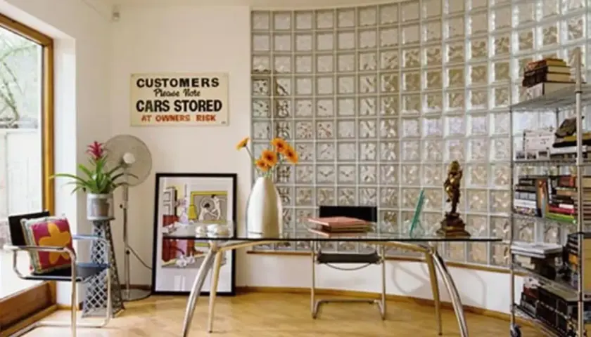 Un ufficio moderno con una parete curva in vetrocemento, scrivania in vetro, sedia, ventilatore e scaffali pieni di libri.