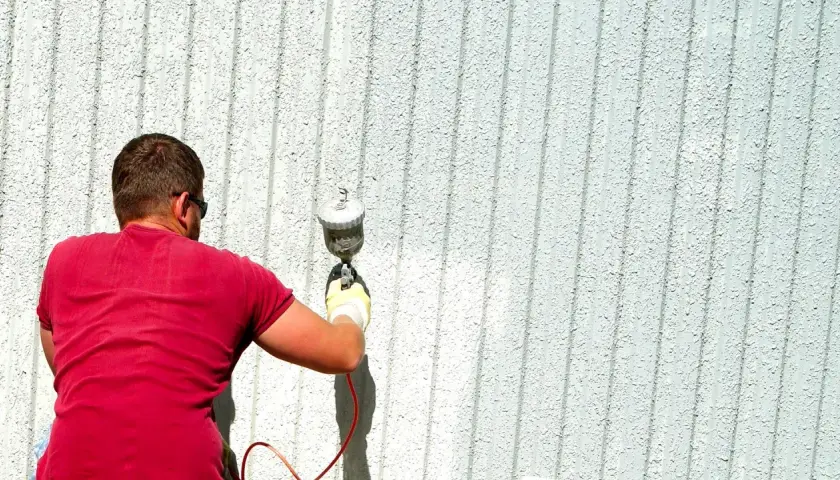 Uomo con maglietta rossa e occhiali da sole impara come verniciare a spruzzo una parete esterna con una pistola a spruzzo.