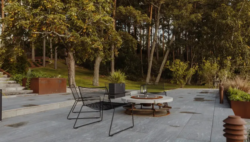 Terrazza con pavimentazione in gres flottante, sedie nere e tavolino con braciere, circondata da alberi e vegetazione.