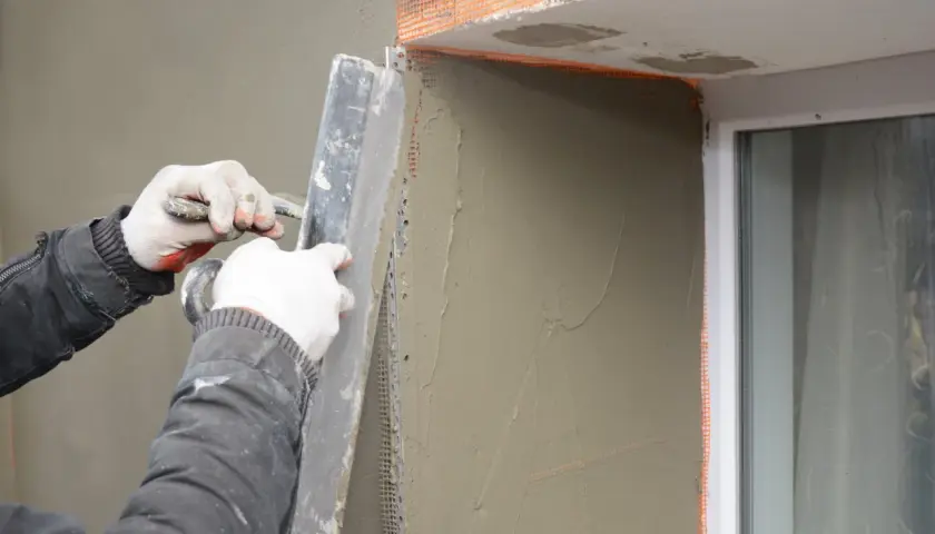 Lavoratore applica intonaco alle spallette di finestre esistenti, usando una cazzuola e rete di rinforzo.