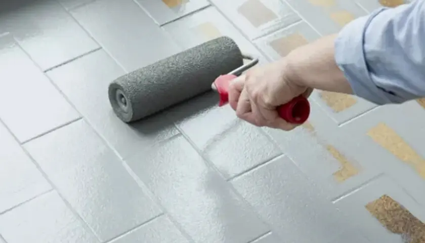 Applicazione di smalto per pavimenti: pro e contro di un restyling fai-da-te. Un rullo grigio stende uniformemente la vernice su piastrelle grigie.
