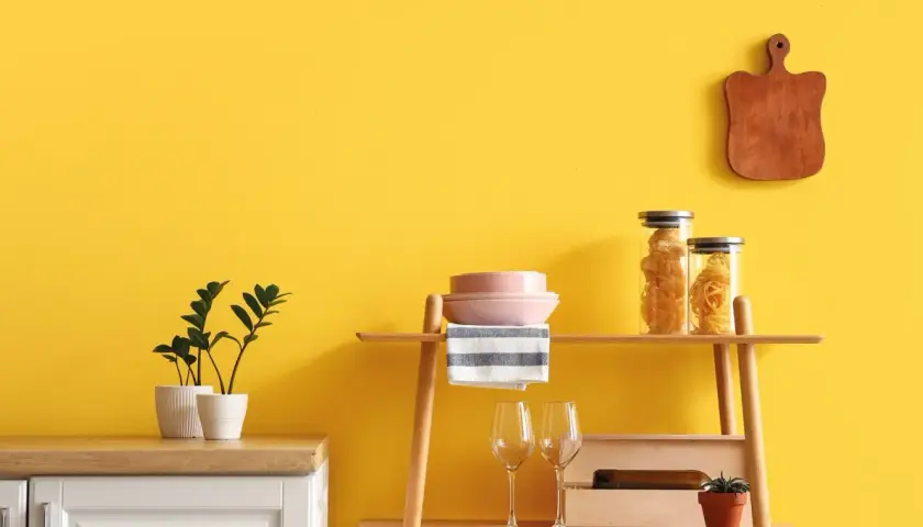 Cucina con pareti giallo vivace, dove **vernici naturali** esaltano gli arredi: piante, pasta in barattoli, piatti rosa e un tagliere in legno.
