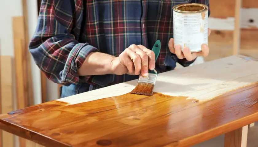Mano che stende flatting su un piano in legno grezzo con un pennello, dando una finitura lucida e protettiva.