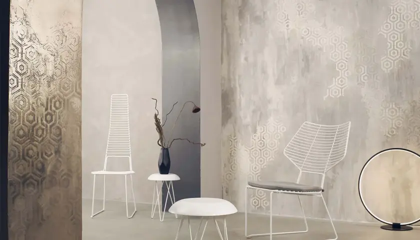 Parete con texture materica e motivi esagonali, che mostra diversi tipi di pittura decorativa. Due sedie bianche minimaliste e tavolini completano l'arredo.