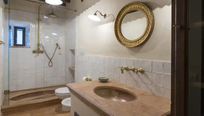 Bagno con piastrelle bianche quadrate, lavabo in cotto, specchio dorato e doccia con rubinetteria in ottone.