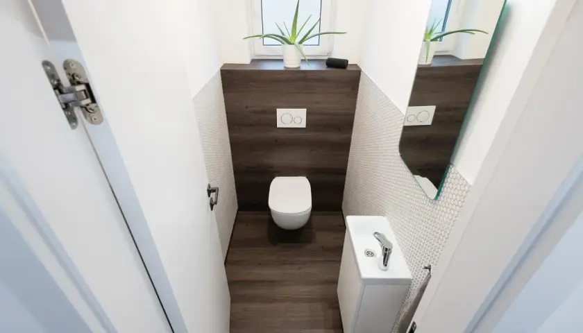 Bagno piccolo stretto con WC, lavandino bianco, specchio e pianta. Pareti con piastrelle bianche a mosaico e inserto in legno scuro.
