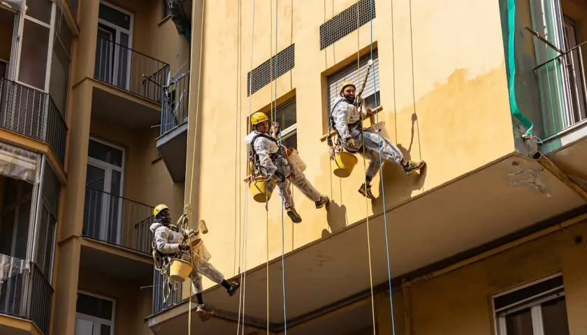 Operai in corda applicano il miglior intonaco per esterni su un edificio giallo. Lavori in quota per un risultato impeccabile.