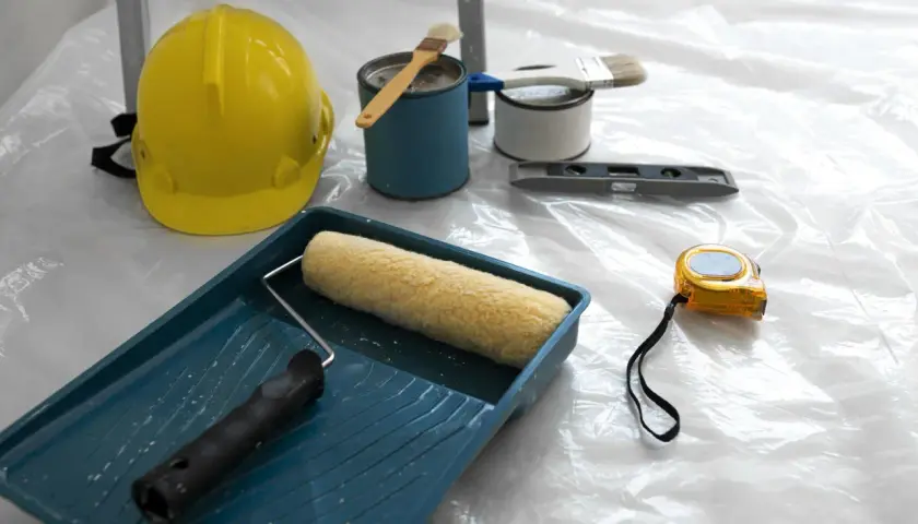 Attrezzi da lavoro: casco giallo, rullo, pennelli, livella, metro e barattoli di vernice, pronti per applicare l'intonaco per umidità.
