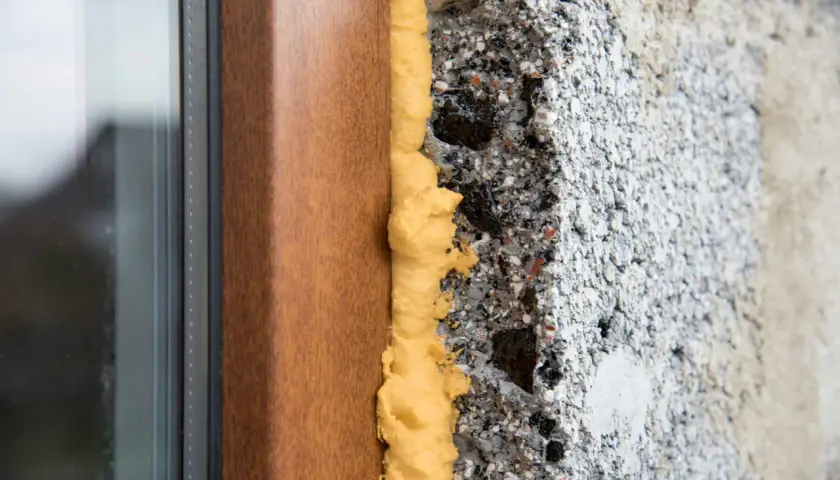 Schiume poliuretaniche espandenti sigillano il vano tra un telaio di finestra in legno e un muro grezzo, garantendo isolamento termico.