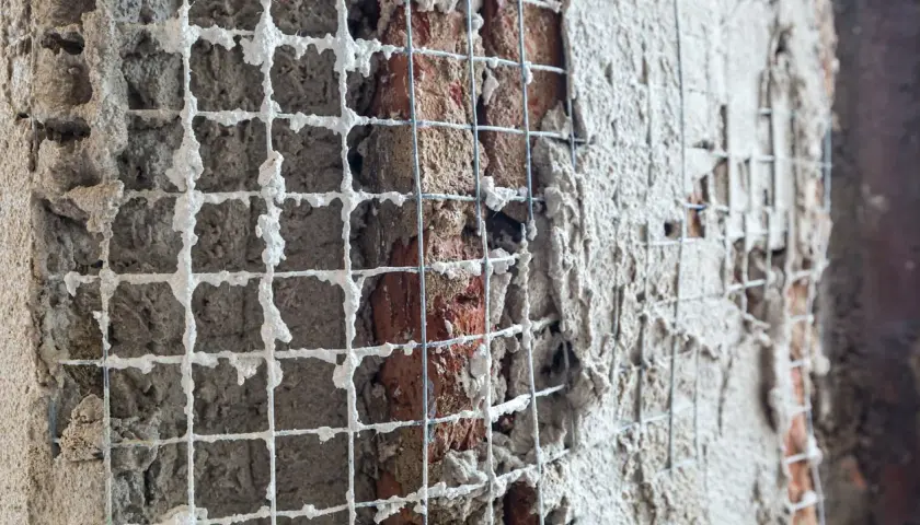 Rete metallica applicata a un muro di mattoni per un rinforzo strutturale, con intonaco fresco che la ricopre parzialmente.