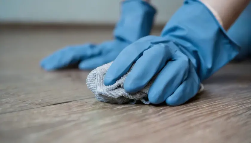 Mani con guanti blu puliscono un pavimento in legno con un panno, mostrando come togliere il miele dal pavimento.