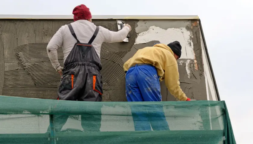 Lavoratori applicano intonaco su un muro esterno, integrando una rete per rinforzo. La rasatura del muro esterno con rete è in corso.