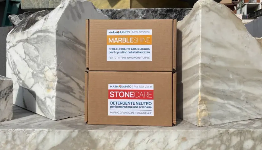 Due scatole di prodotti per la pulizia del marmo, "MarbleShine" e "Stonecare", sono posizionate tra blocchi di marmo grezzo. Ideale per sapere come lavare pavimento in marmo.