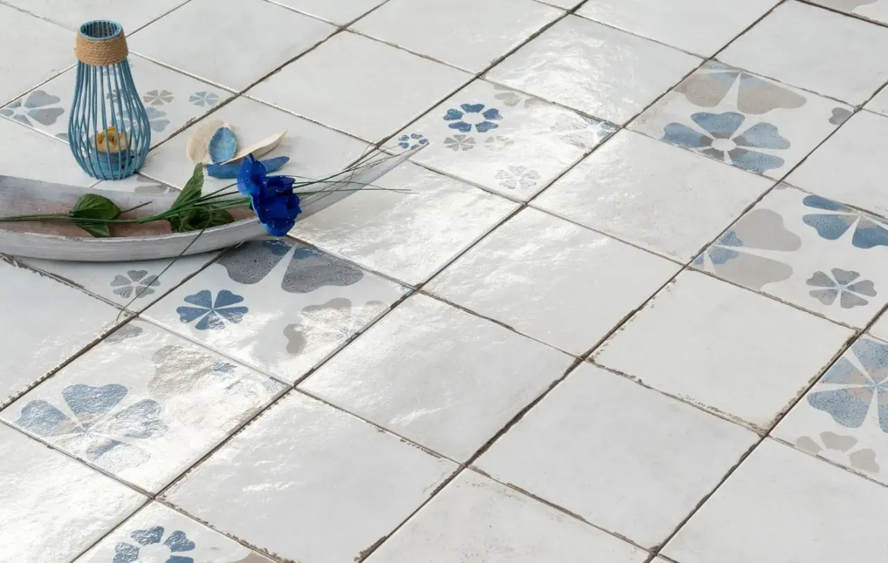 Pavimento con piastrelle quadrate, alcune decorate con fiori blu e grigi, altre bianche. Un vassoio in legno con un fiore blu e un portacandele blu completano la scena, mostrando la differenza tra mattonella e piastrella.