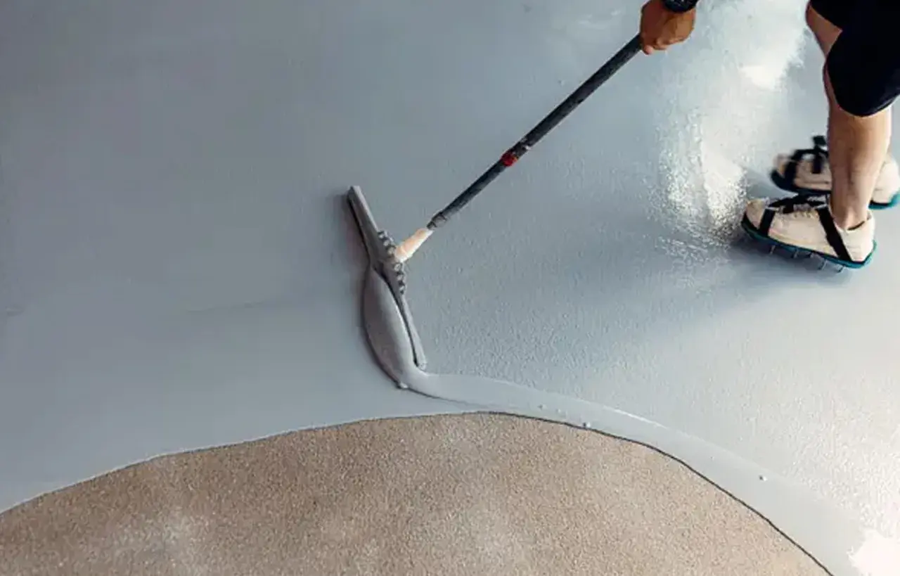 Applicazione di resina per un pavimento fai da te. Una spatola stende il materiale su una superficie grigia, mentre una persona con scarpe chiodate cammina sul cemento grezzo.