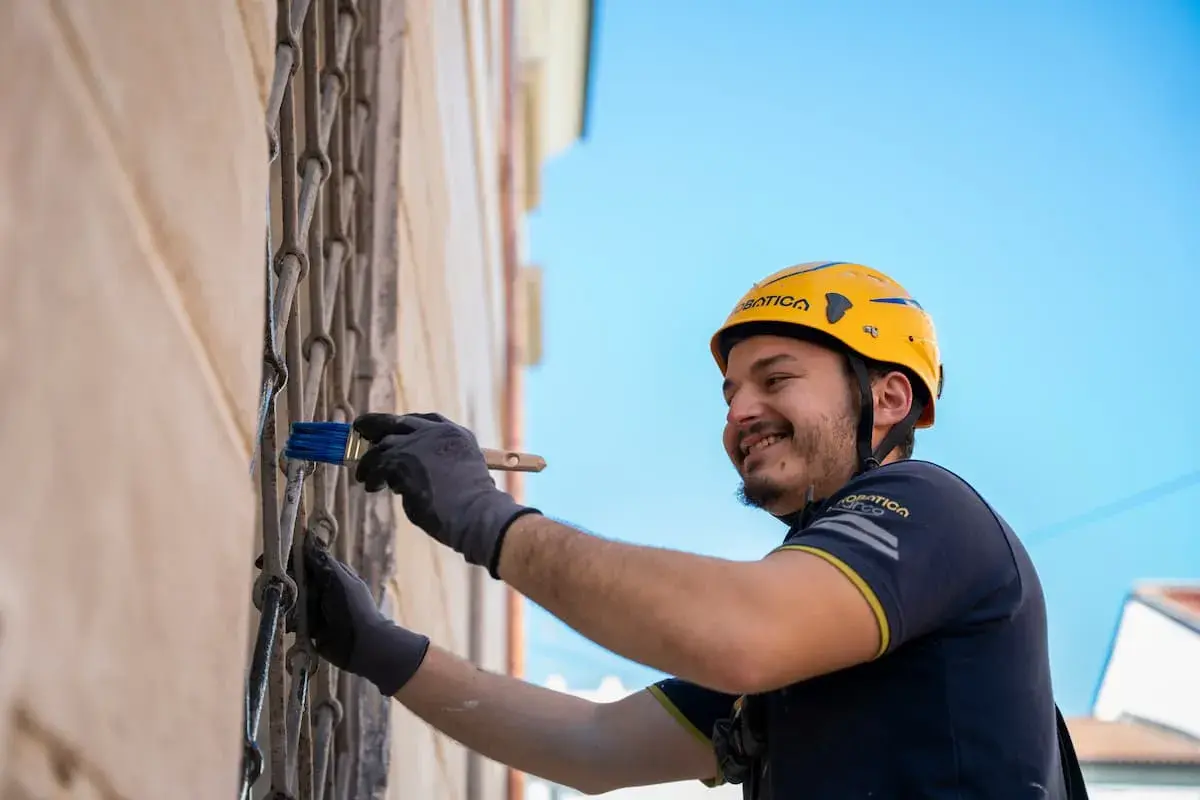 Operaio con casco giallo applica pittura su grata metallica, parte di un sistema di intonaco armato con rete in fibra di vetro.