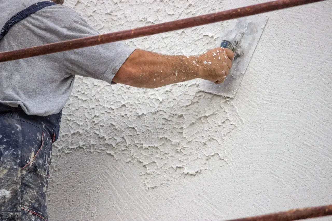 Un operaio applica intonaco rustico con una spatola, creando una texture grezza e irregolare su una parete esterna.