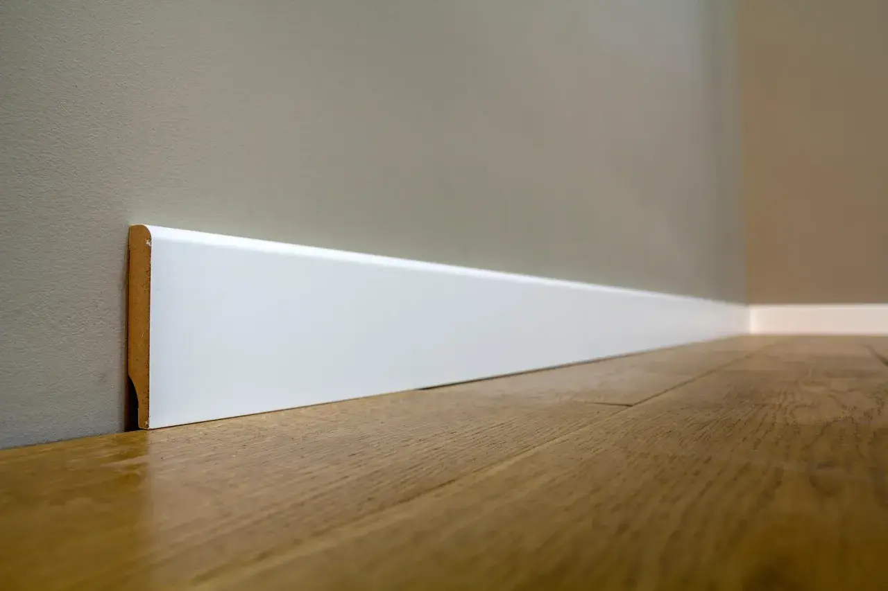Battiscopa bianco moderno, con un'altezza che definisce lo spazio. Il legno del pavimento riflette la luce.