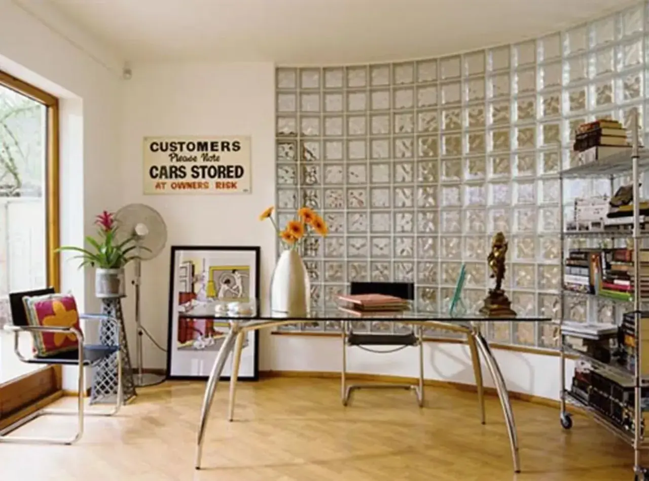 Un ufficio moderno con una parete curva in vetrocemento, scrivania in vetro, sedia, ventilatore e scaffali pieni di libri.