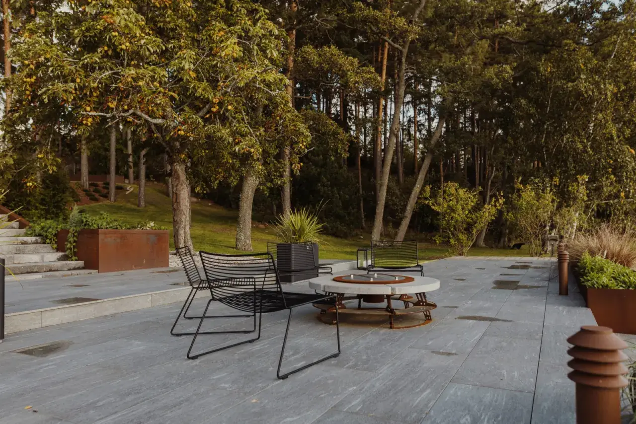 Terrazza con pavimentazione in gres flottante, sedie nere e tavolino con braciere, circondata da alberi e vegetazione.