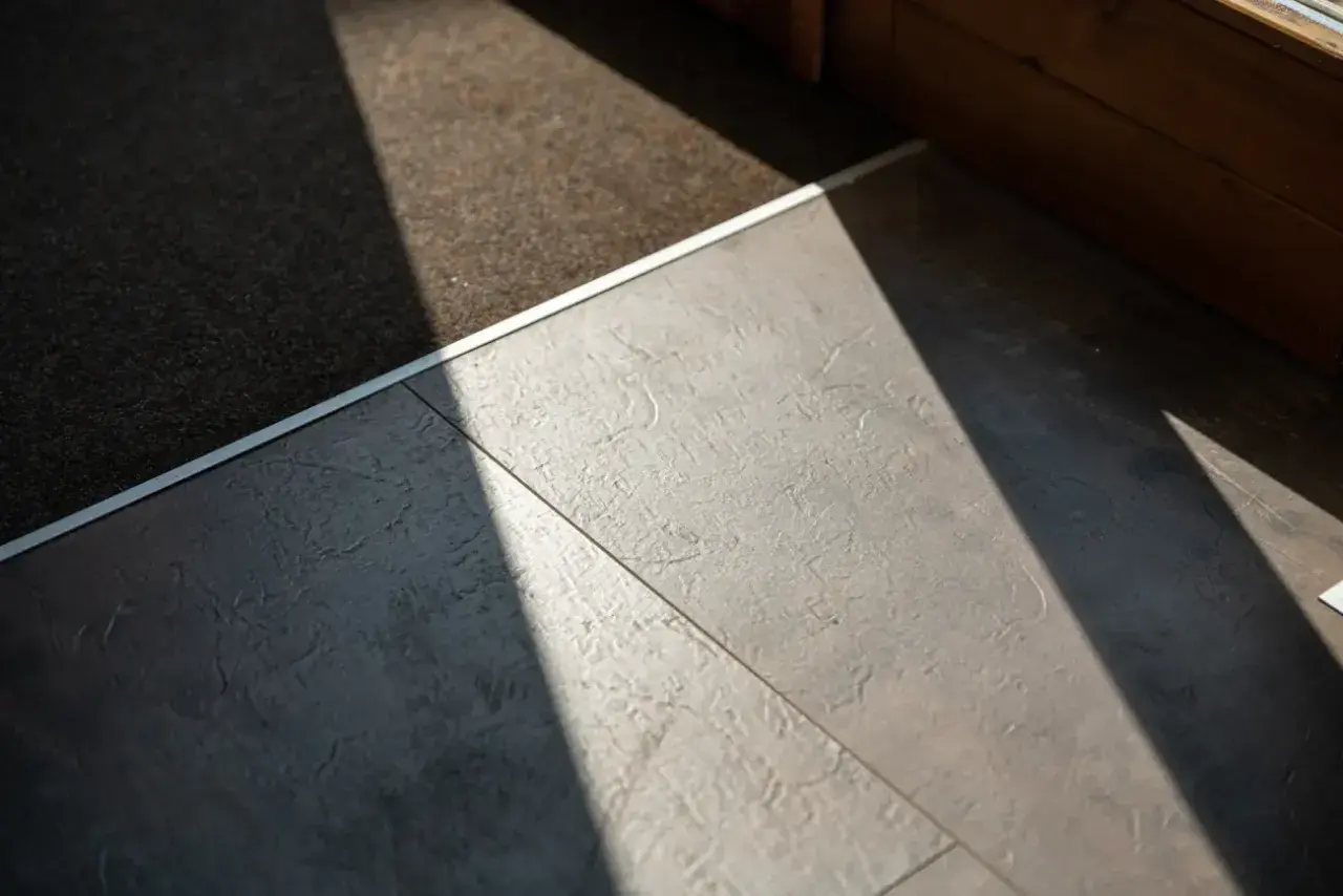 Un tappeto scuro incontra un moderno pavimento flottante grigio, illuminato da un raggio di sole diagonale.