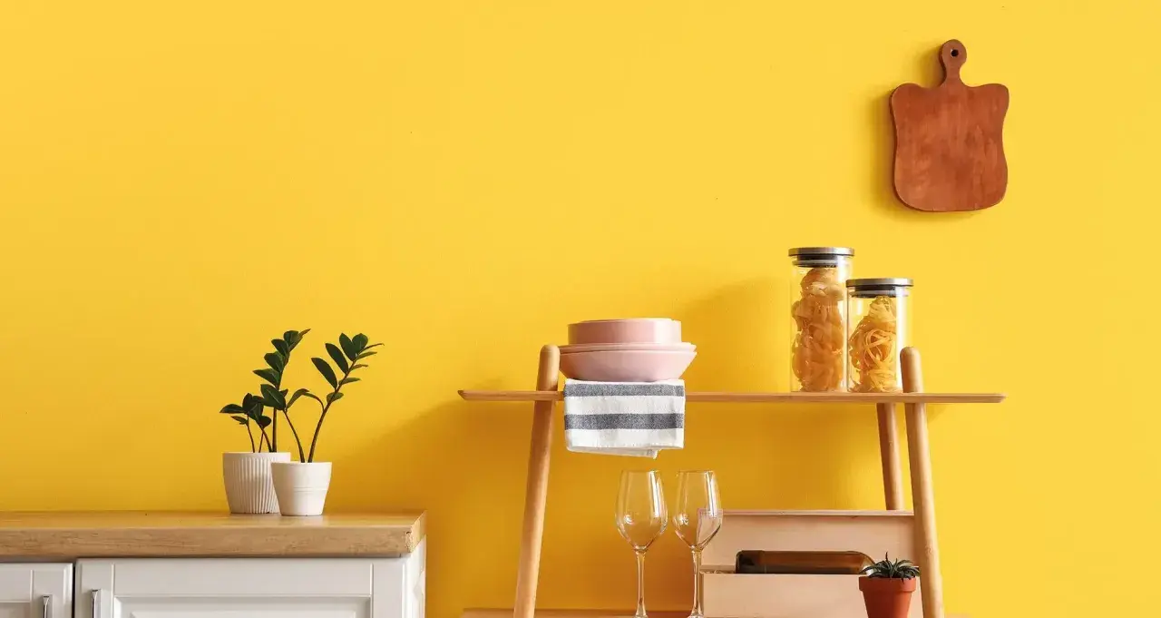 Cucina con pareti giallo vivace, dove **vernici naturali** esaltano gli arredi: piante, pasta in barattoli, piatti rosa e un tagliere in legno.