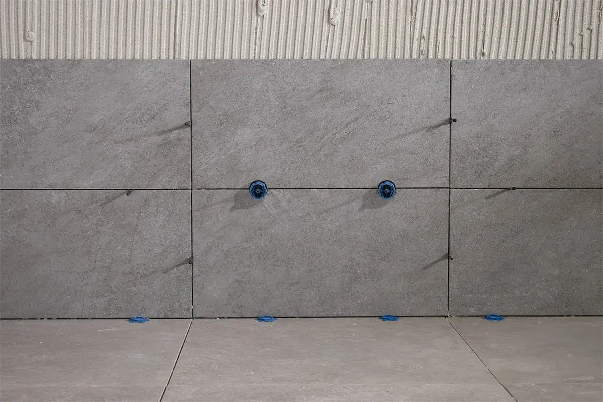 Posa di piastrelle grigie con distanziatori blu. Le fughe piastrelle non rettificate sono visibili, con adesivo sottostante.