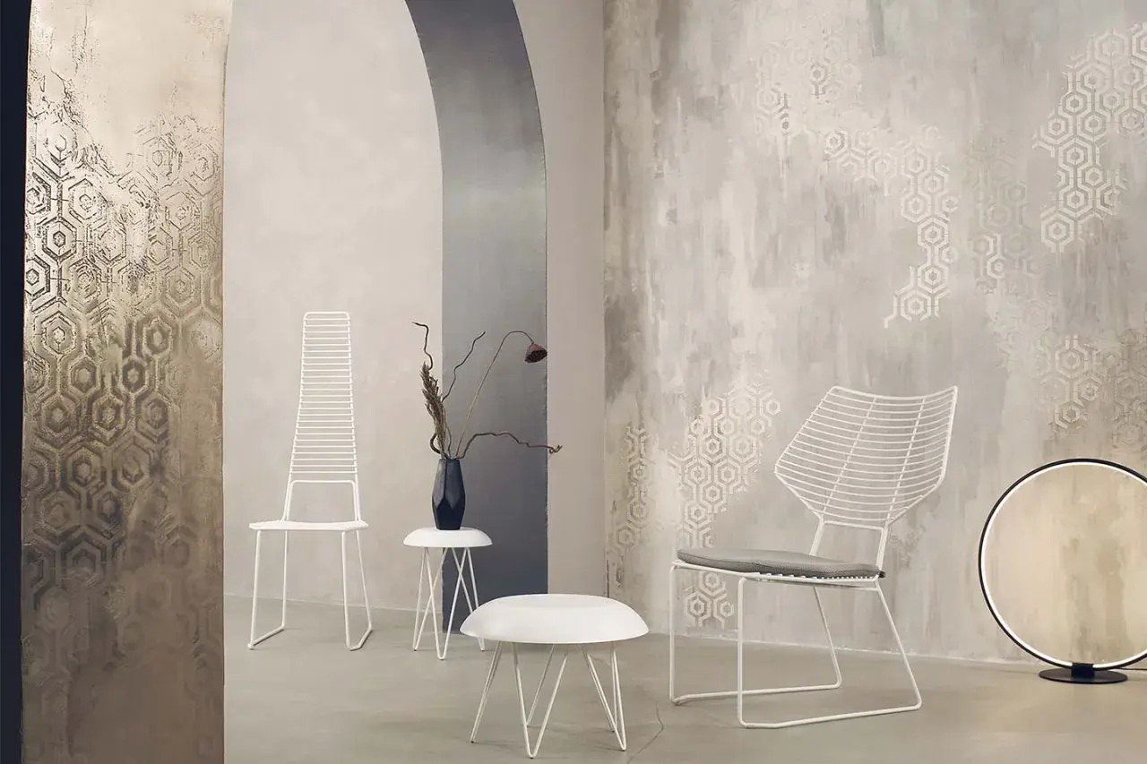 Parete con texture materica e motivi esagonali, che mostra diversi tipi di pittura decorativa. Due sedie bianche minimaliste e tavolini completano l'arredo.