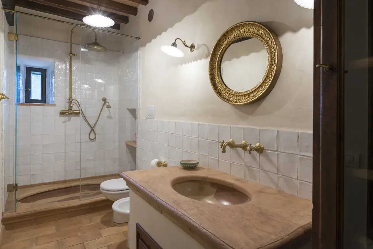 Bagno con piastrelle bianche quadrate, lavabo in cotto, specchio dorato e doccia con rubinetteria in ottone.