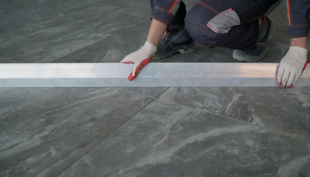 Mano con guanto bianco e rosso posiziona un profilo metallico su piastrelle grigie 60x120. Preventivo prezzo posa piastrelle 60x120.