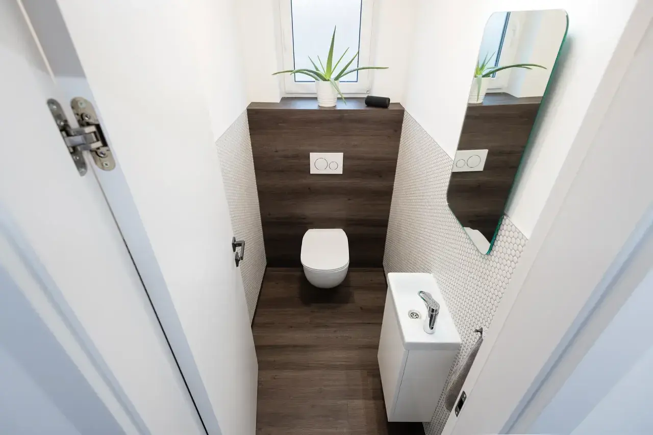 Bagno piccolo stretto con WC, lavandino bianco, specchio e pianta. Pareti con piastrelle bianche a mosaico e inserto in legno scuro.
