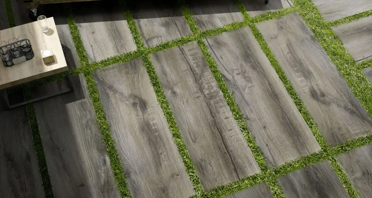 Pavimento posa a secco su erba, con doghe effetto legno grigio alternate a strisce di prato verde. Un tavolino da caffè completa la scena.