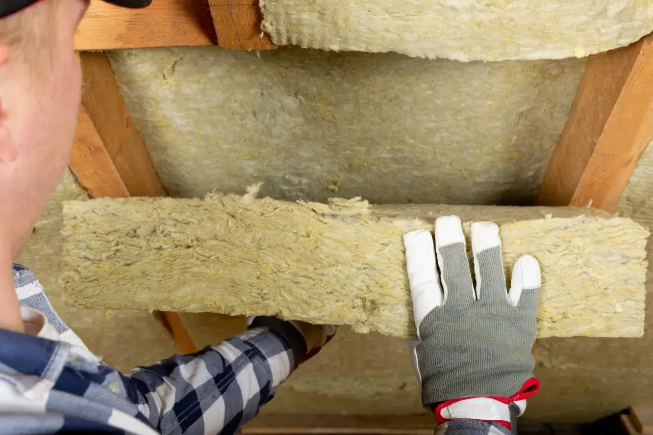 Operaio con guanti installa pannelli di lana di roccia isolante tra le travi di legno di un sottotetto.