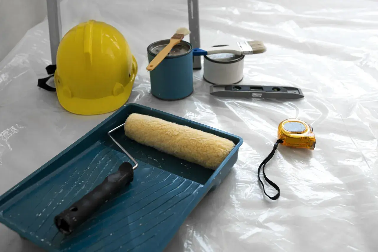 Attrezzi da lavoro: casco giallo, rullo, pennelli, livella, metro e barattoli di vernice, pronti per applicare l'intonaco per umidità.