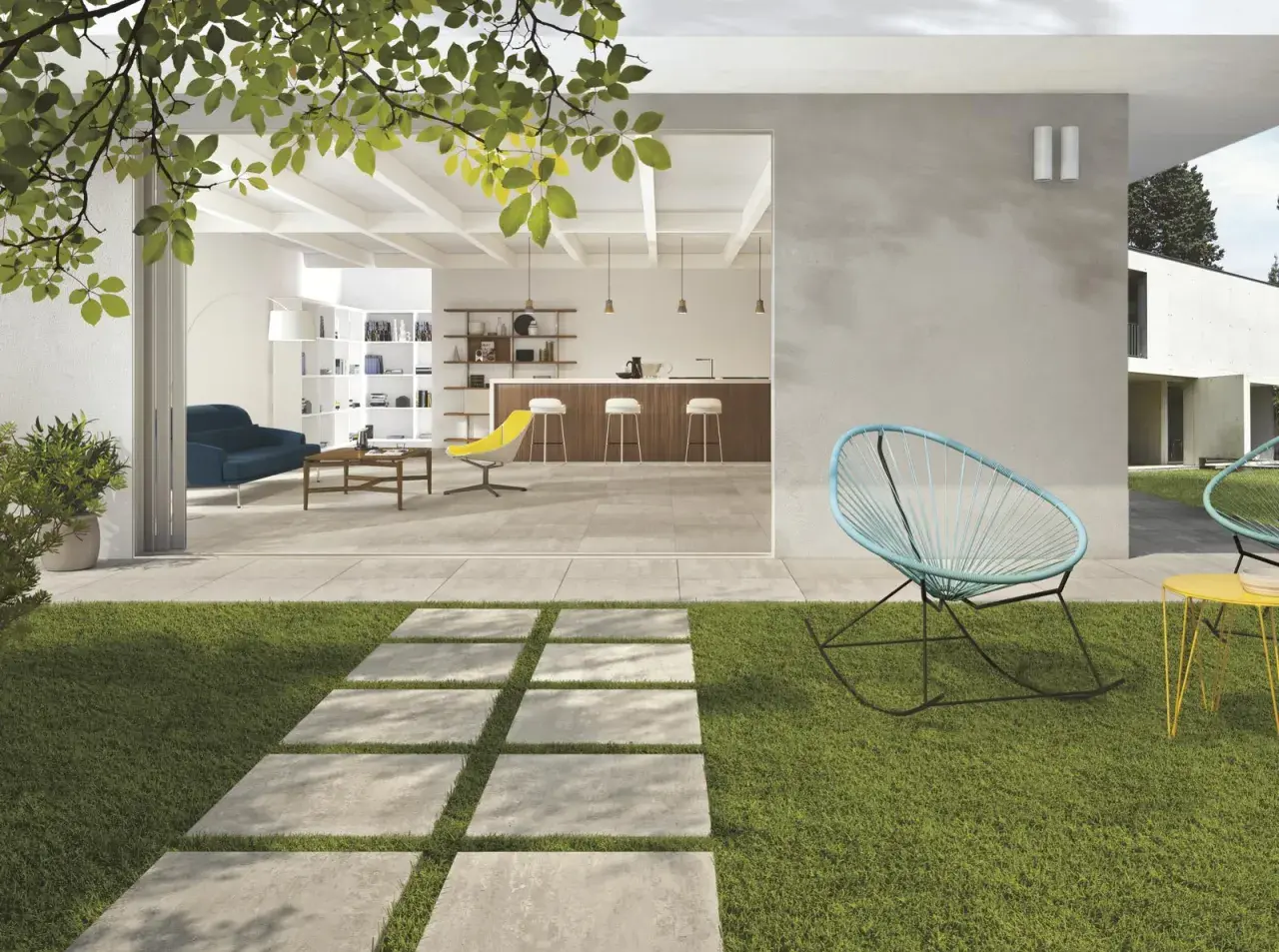 Un sentiero in lastre quadrate su prato verde conduce a un'area living con pavimentazione esterna moderna, che si apre su un interno luminoso con cucina e soggiorno.