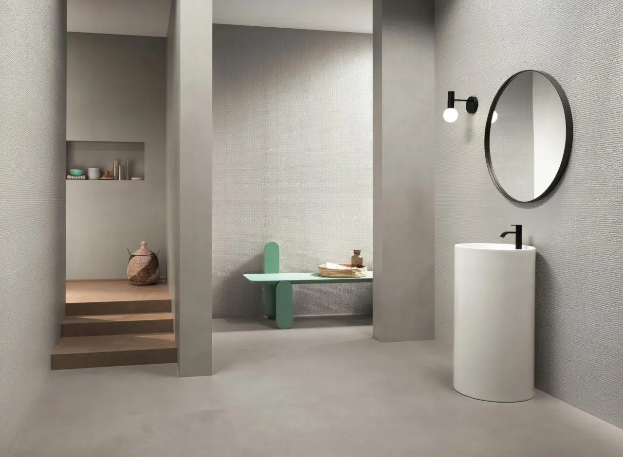 Bagno moderno con rivestimento uguale al pavimento, lavabo a colonna, specchio rotondo e nicchia con oggetti decorativi.