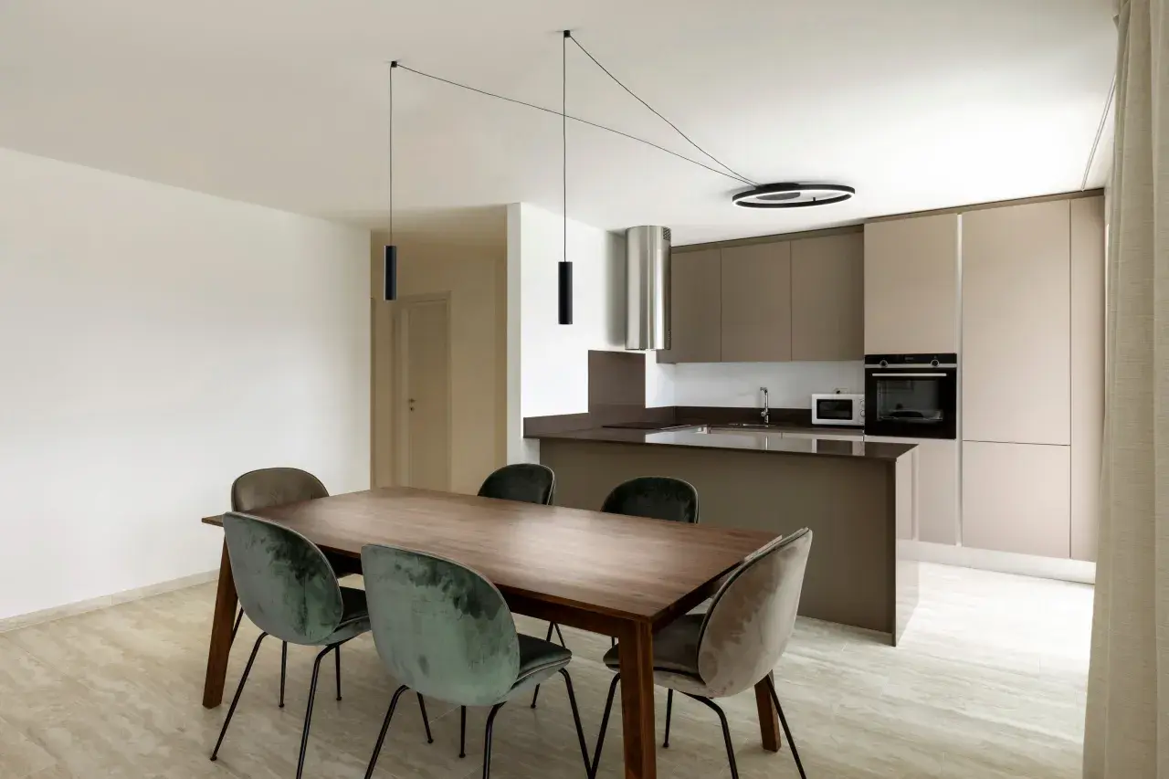 Un lampadario decentrato crea un'atmosfera moderna sopra un tavolo da pranzo in legno con sedie in velluto verde. Soluzioni di illuminazione eleganti per ogni spazio.