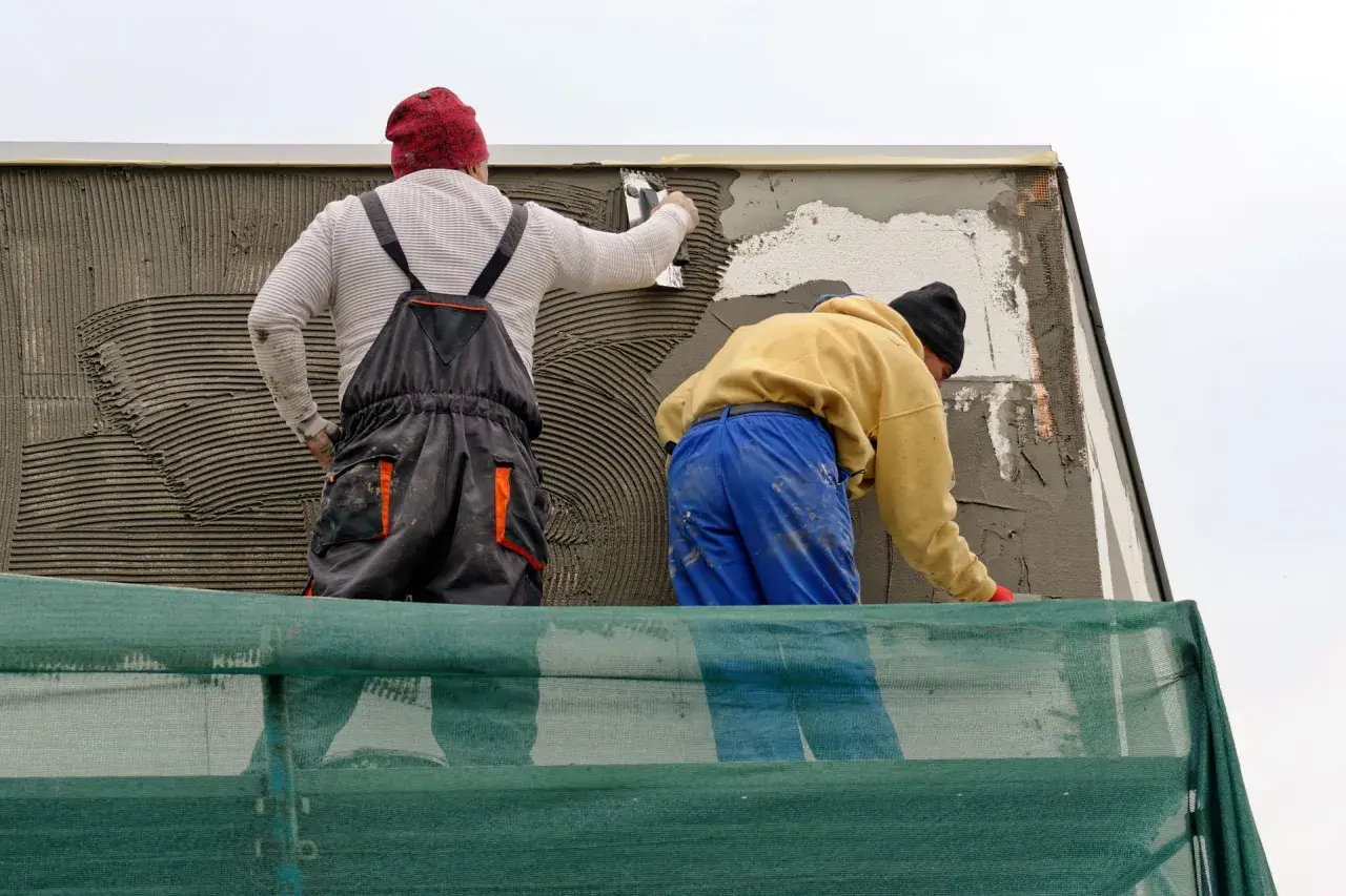 Lavoratori applicano intonaco su un muro esterno, integrando una rete per rinforzo. La rasatura del muro esterno con rete è in corso.