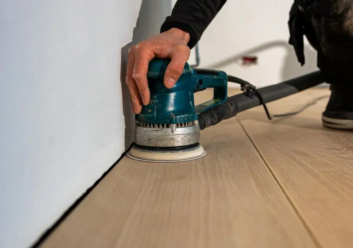 Mano che usa una levigatrice orbitale per levigare il parquet fai da te, avvicinandosi al battiscopa bianco.