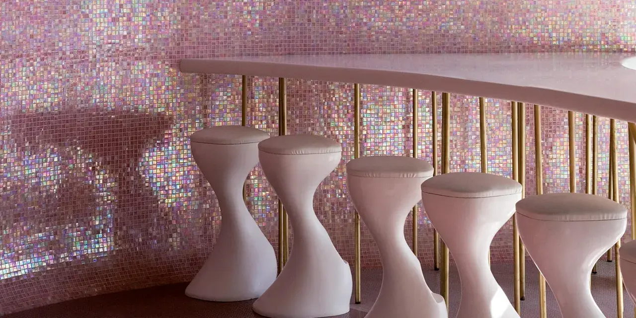 Un bancone rosa pallido con sgabelli bianchi si staglia contro un muro scintillante di **bisazza ceramiche** iridescenti, creando un'atmosfera glamour.
