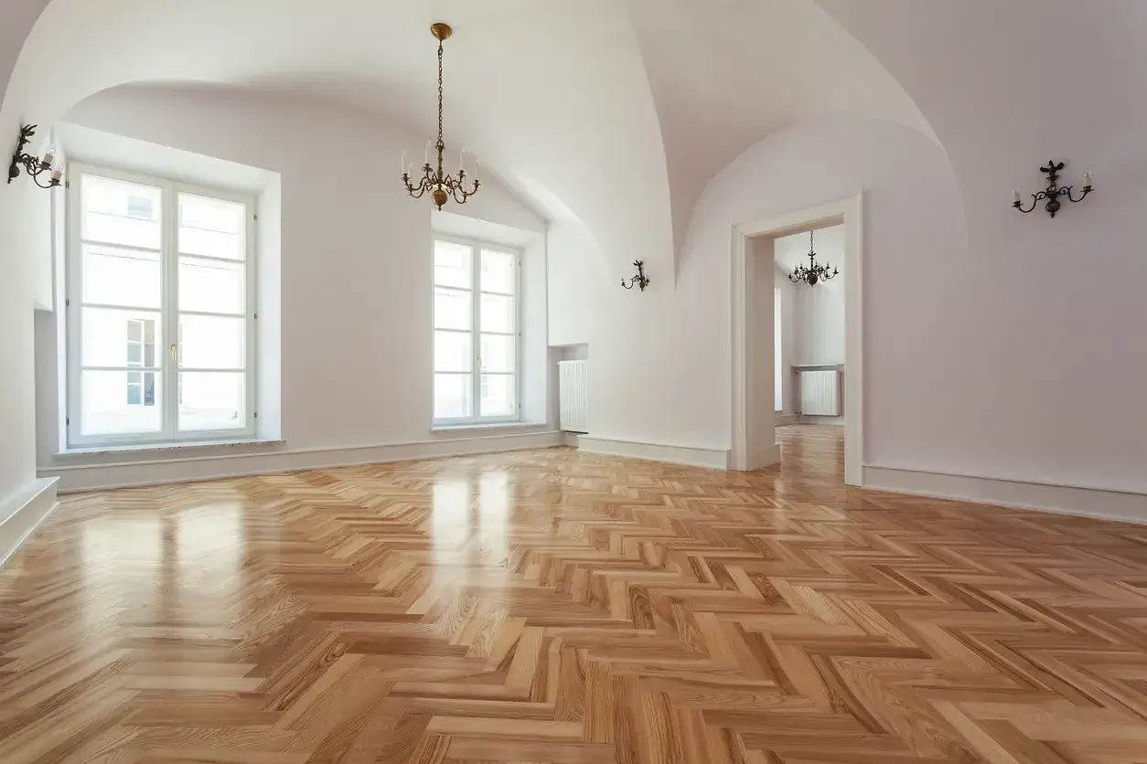 Un parquet a spina di pesce lucido, pronto per essere levigato fai da te. L'ambiente è luminoso, con grandi finestre e lampadari antichi.