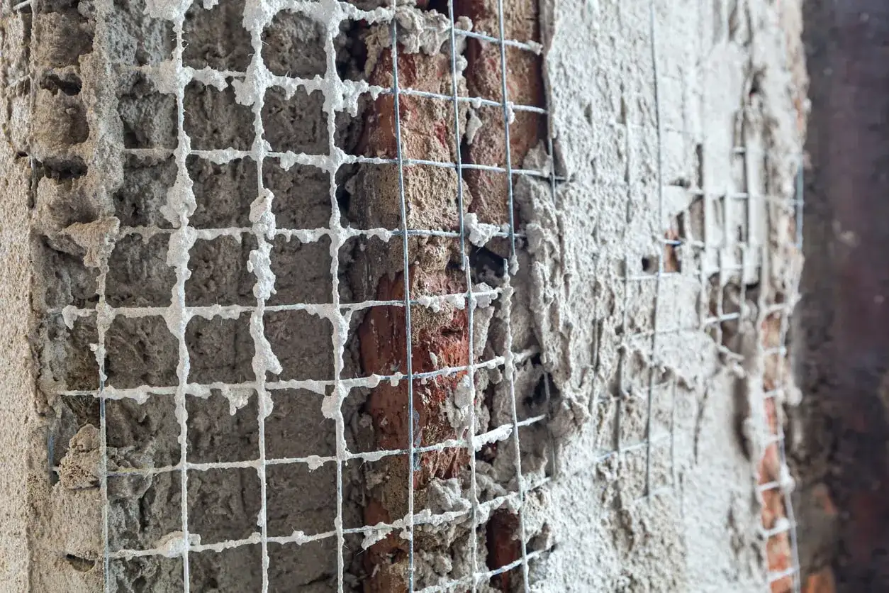 Rete metallica applicata a un muro di mattoni per un rinforzo strutturale, con intonaco fresco che la ricopre parzialmente.