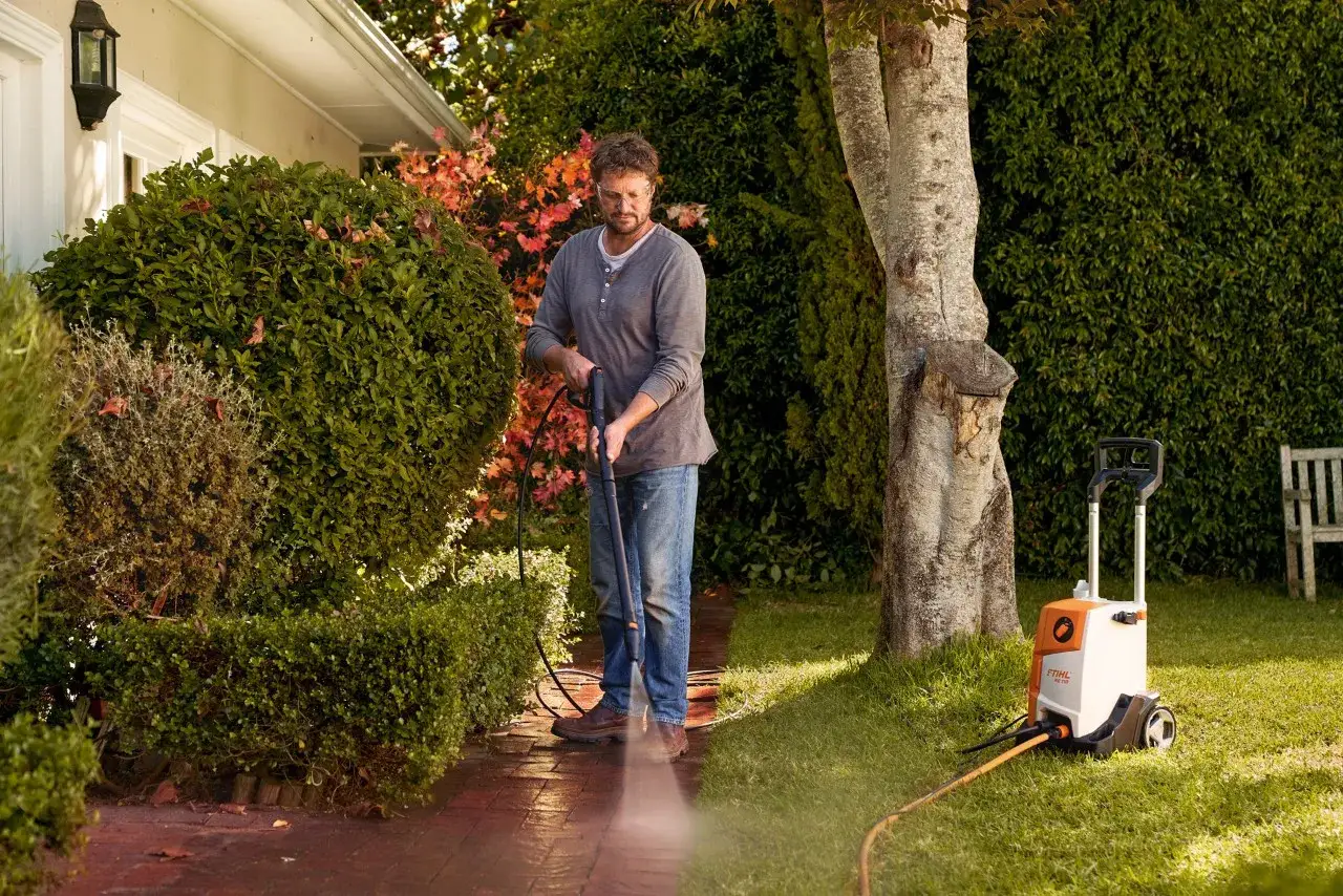 Man pressure washing a brick pathway next to lush green bushes and a tree.