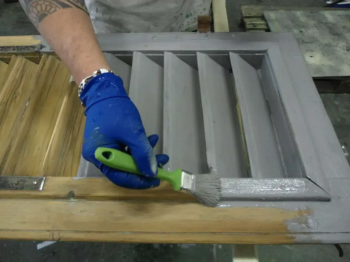 Mano con guanto blu applica vernice per persiane in legno con pennello verde.
