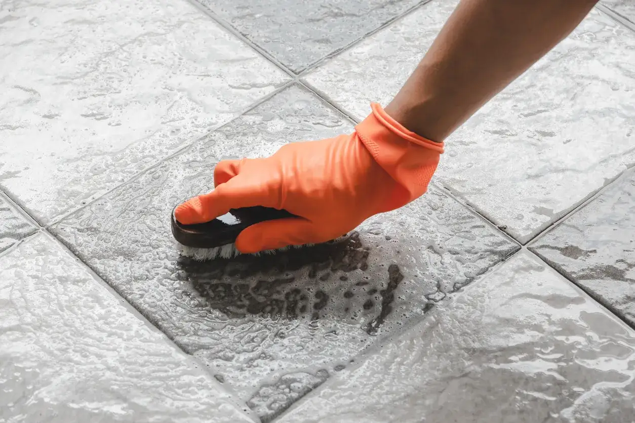 Mano con guanto arancione che pulisce le piastrelle del bagno con una spazzola e schiuma.