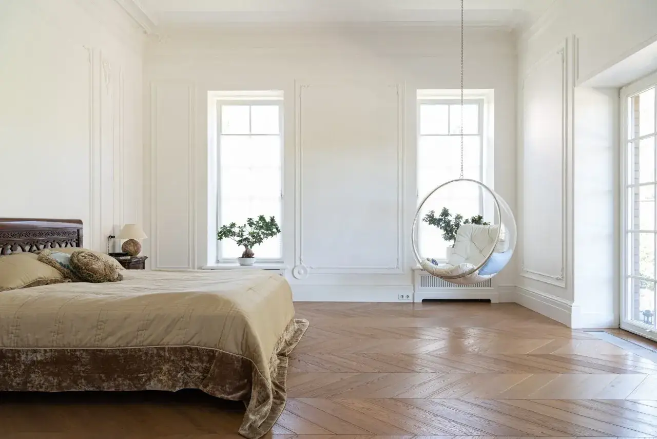 Camera da letto luminosa con letto, sedia sospesa e un elegante pavimento a spina di pesce che riflette la luce.