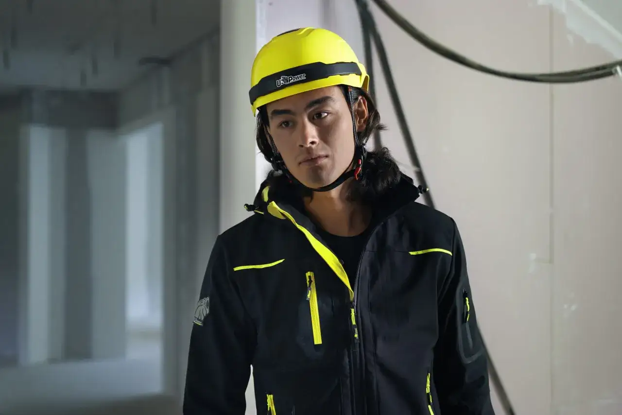 Giovane operaio con elmetto da cantiere normativa, indossa giacca nera con dettagli gialli.