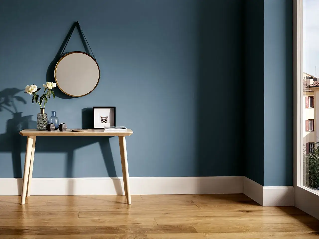Un tavolino di legno con fiori e un quadro di procione, appoggiato a una parete blu. L'altezza battiscopa bianco risalta sul pavimento in legno.