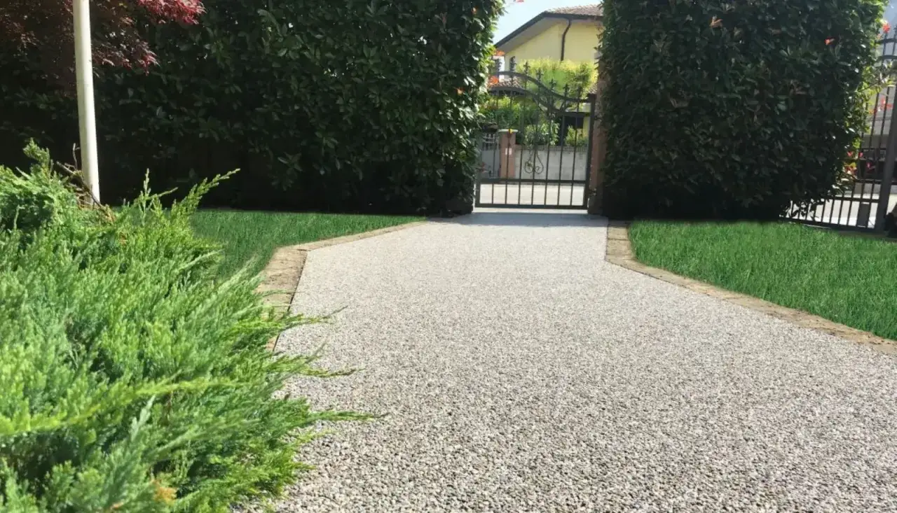 Un vialetto carrabile economico con ghiaia resinata, bordato da siepi verdi e prato curato, conduce a un cancello in ferro battuto.